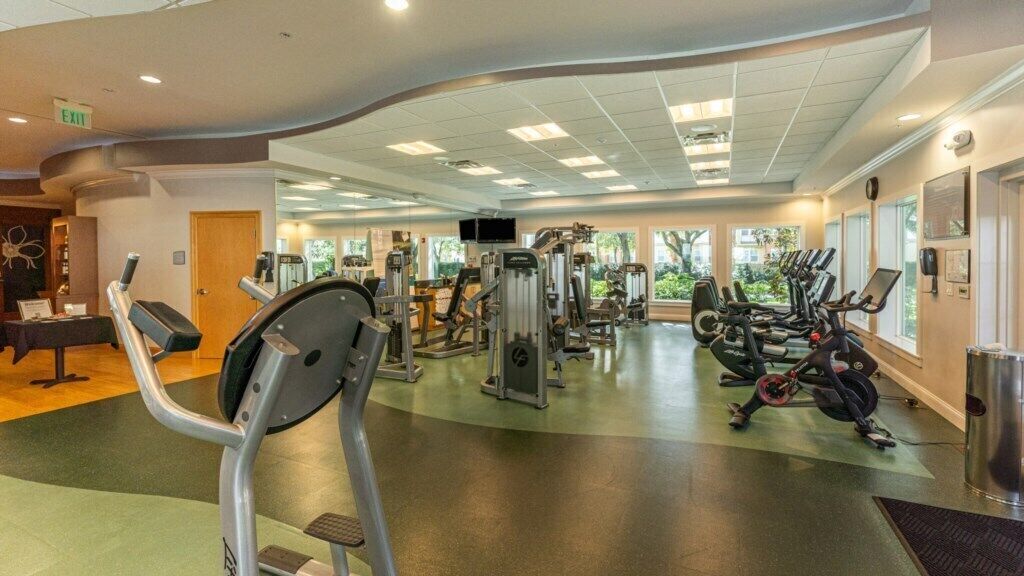 Une salle de sport équipée de divers appareils d’exercice, un comptoir d’accueil et de grandes fenêtres.