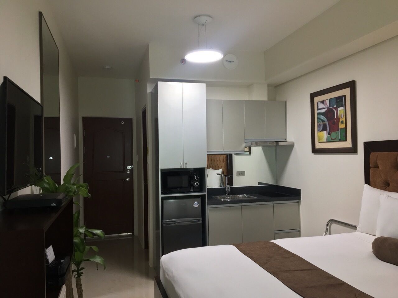 Ein kompaktes Hotelzimmer mit Bett, einer kleinen Küchenzeile, einem Flachbildfernseher und einem Bild an der Wand.