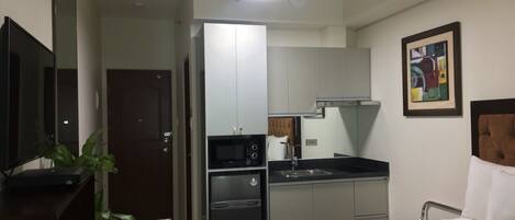 A compact hotel room with a bed, a small kitchenette, a flat-screen TV, and a painting on the wall.