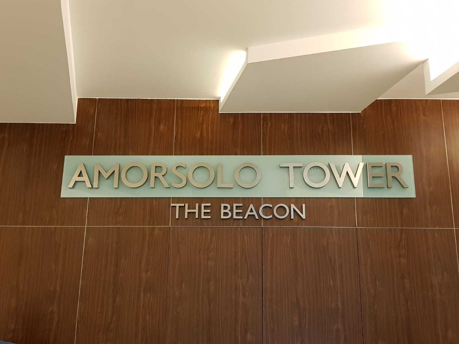 A sign for Amorsolo Tower, The Beacon, with a wooden panel background and a light fixture above.