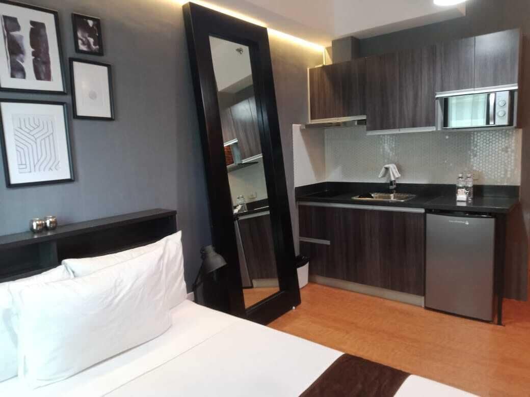 Habitación de hotel compacta con cama, mesita de noche, espejo, kitchenette pequeña con microondas y lavavajillas, y cuadros en la pared.