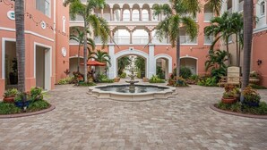 A courtyard with a central fountain, surrounded by palm trees and potted plants, with a multi-story building featuring balconies in the background.