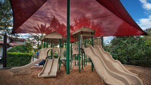 A playground with slides and climbing structures, surrounded by trees and covered by a large red canopy.