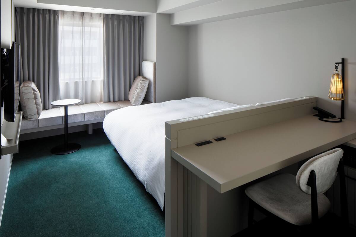A hotel room with a bed, a desk, a chair, a small table, and a lamp.