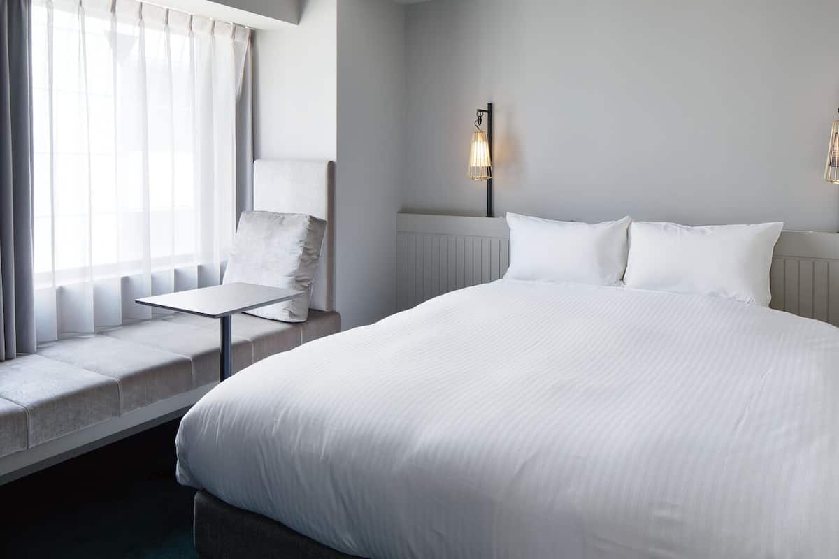 A hotel room with a large bed, a small table, a chair, and two wall-mounted lamps.