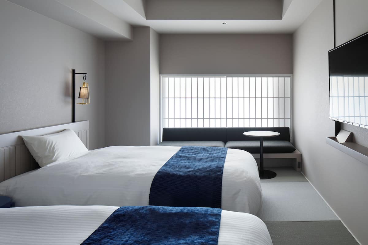 A modern hotel room with a large bed, a small round table, a bench, and a TV mounted on the wall.