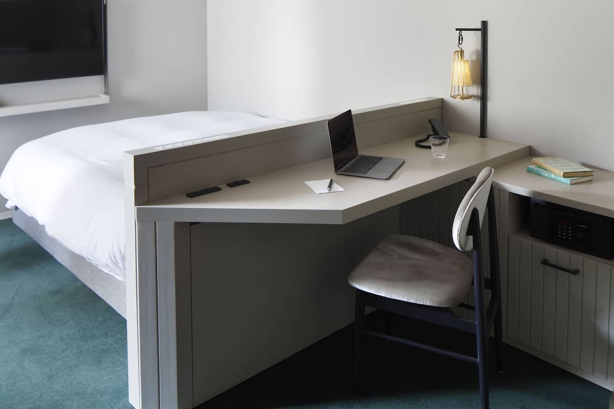 A hotel room with a bed, a desk with a laptop, a chair, a bedside table with a lamp, and a TV mounted on the wall.