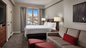 A hotel room with a bed, a TV, a sofa, a red ottoman, and a window with curtains.