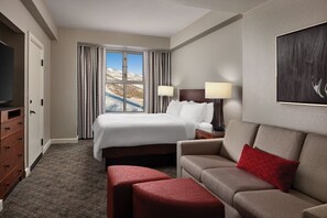 Una habitación de hotel con cama, televisión, sofá, un reposapiés rojo y una ventana con cortinas.