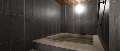 A modern bathroom with dark tiled walls, a wooden ceiling, and a built-in bathtub.