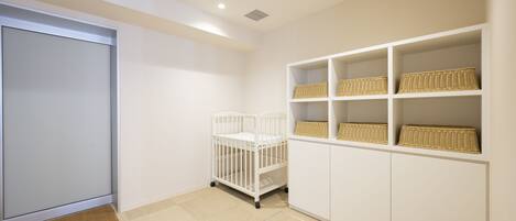 A room with a white crib, white shelving with wicker baskets, and a beige carpet.