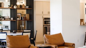 Una sala de estar moderna con sillones de cuero, un sofá y una mesa de centro. Se observa una cocina al fondo con armarios y un refrigerador.