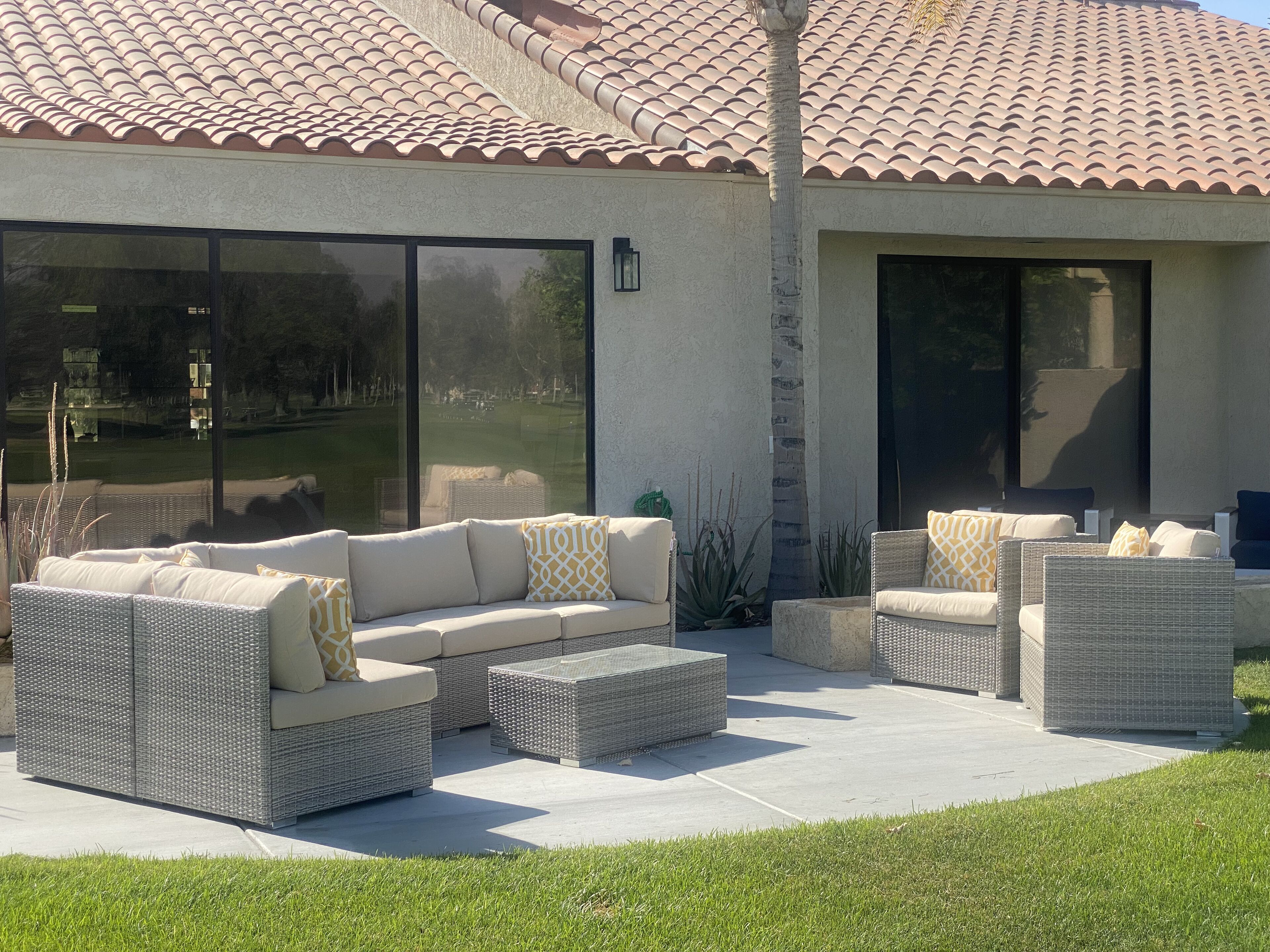 An outdoor patio with wicker furniture, including sofas and ottomans, and a large sliding glass door leading to a grassy area.