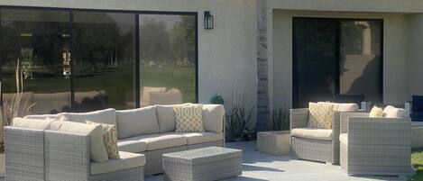An outdoor patio with wicker furniture, including sofas and ottomans, and a large sliding glass door leading to a grassy area.
