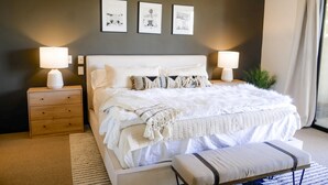 Un dormitorio con una cama grande, dos mesitas de noche con lámparas, un banco y tres cuadros enmarcados en la pared.
