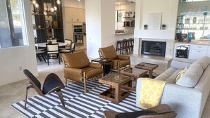 Una sala moderna con un sofá, sillones y una mesa de centro. En el fondo, se observa un comedor con sillas y una mesa.