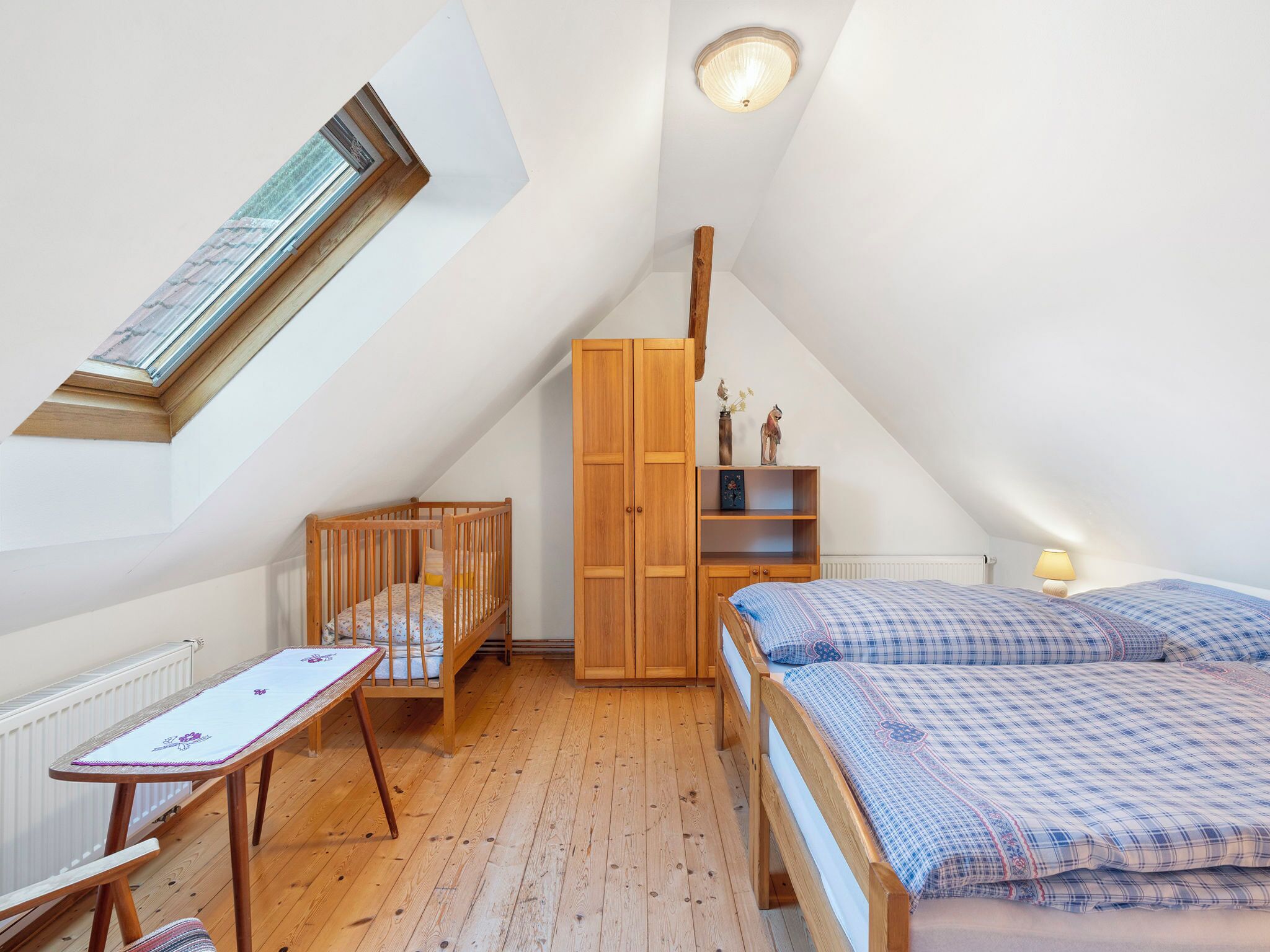 A cozy attic bedroom with a sloped ceiling, a wooden crib, a bed with a checkered duvet, a wooden wardrobe, and a small table with a tablecloth.