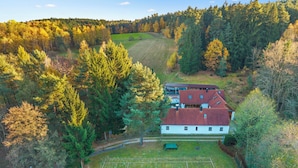 An aerial view of a house with a red roof, surrounded by a tennis court and dense forest.