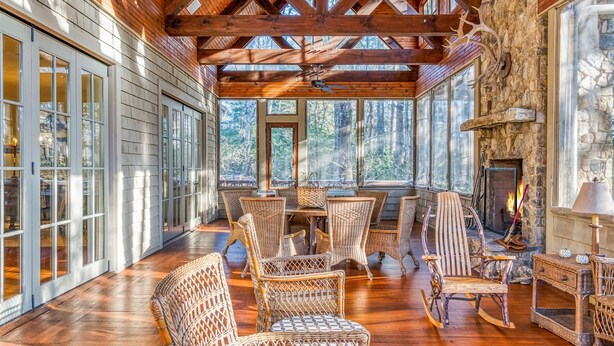 A cozy wooden interior with a stone fireplace, wicker furniture, and large windows offering a forest view. - Rustic Elegance. Three Fireplaces. A Crown Jewel of a Screened Porch | Sagee Woods (Highlands)