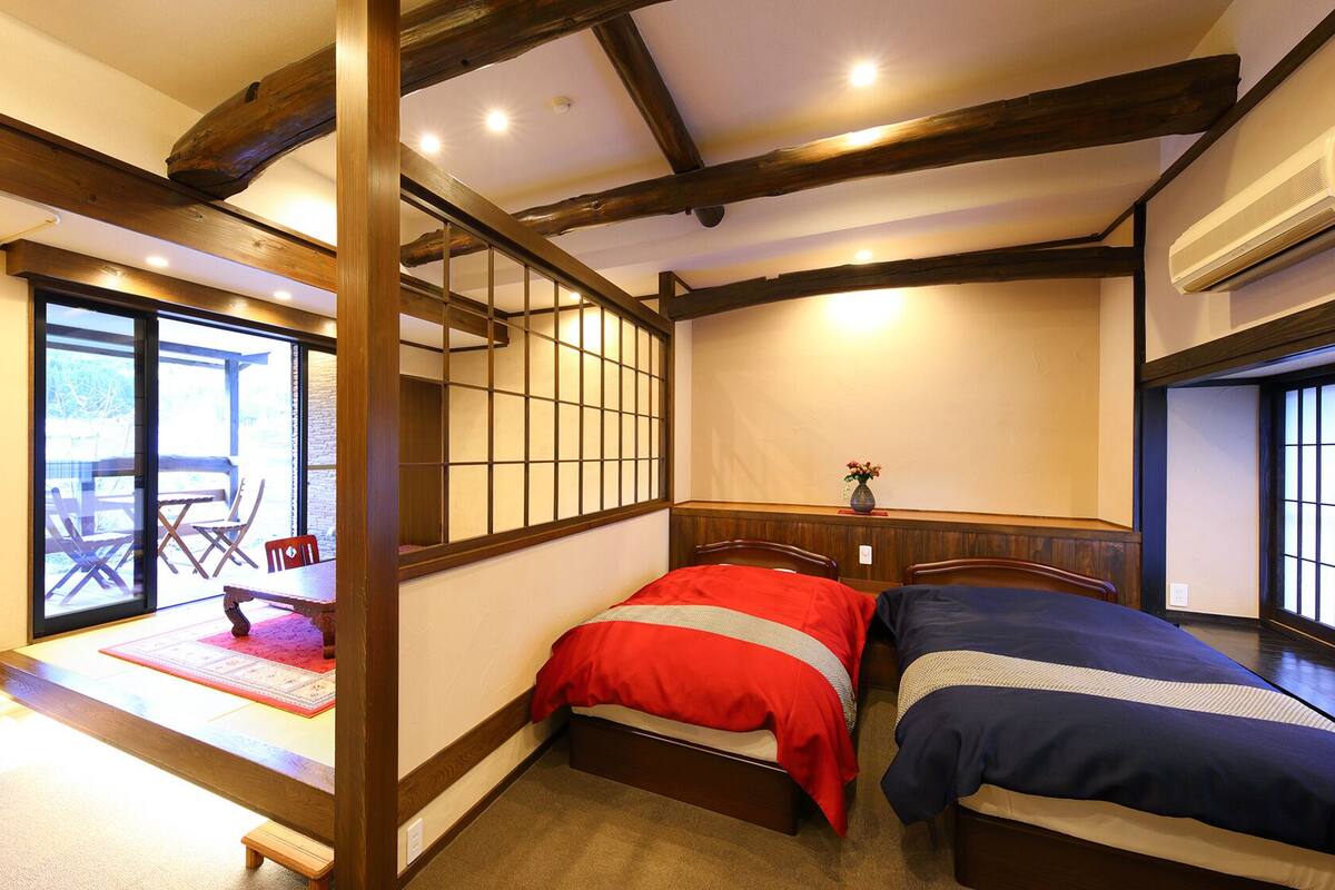 Una habitación tradicional japonesa con suelo de tatami, una cama baja de madera, puertas correderas y vistas a un patio exterior.