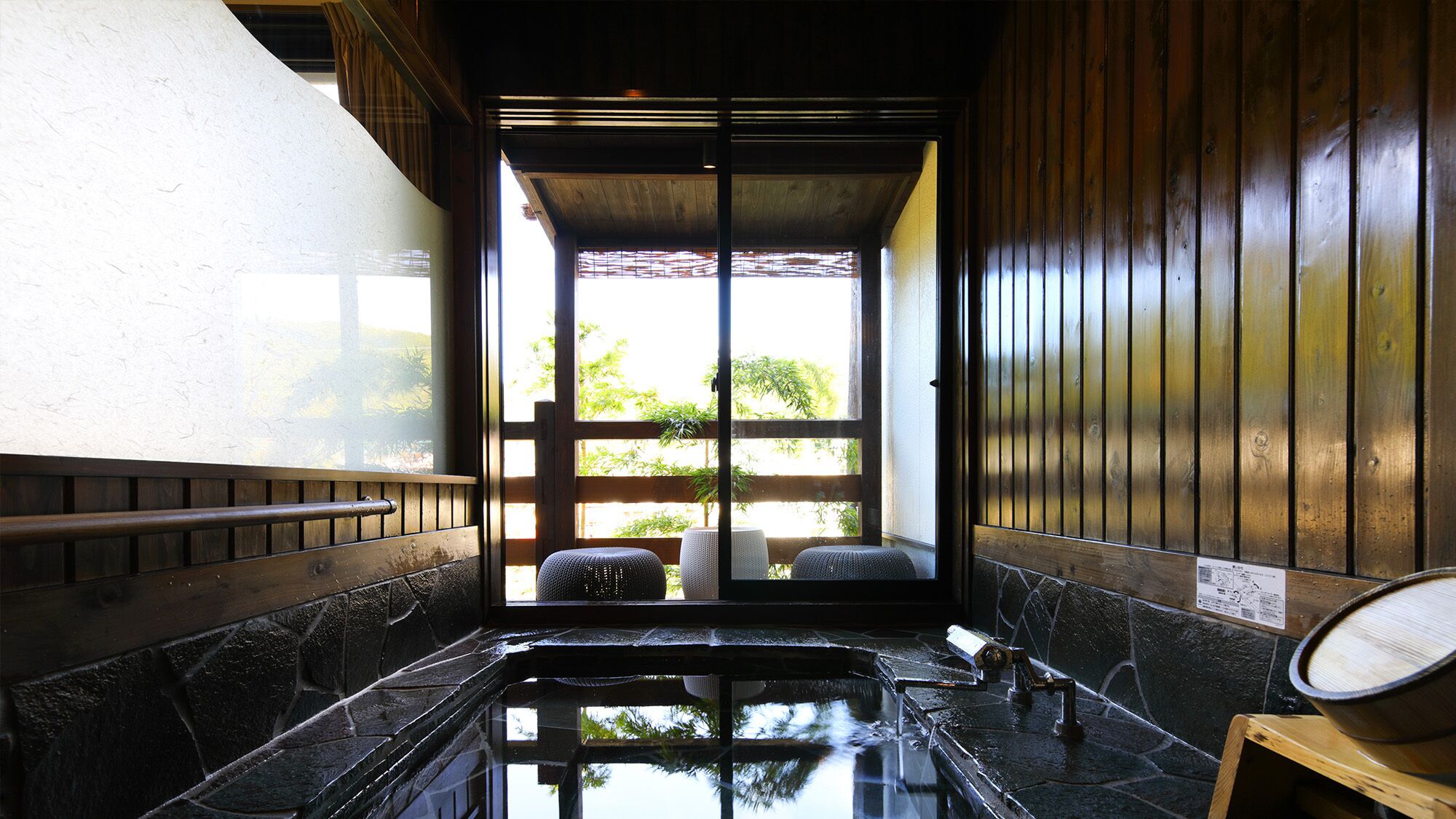 Un jacuzzi con azulejos de piedra, un banco de madera, un taburete pequeño y una mesa de madera con un cuenco.