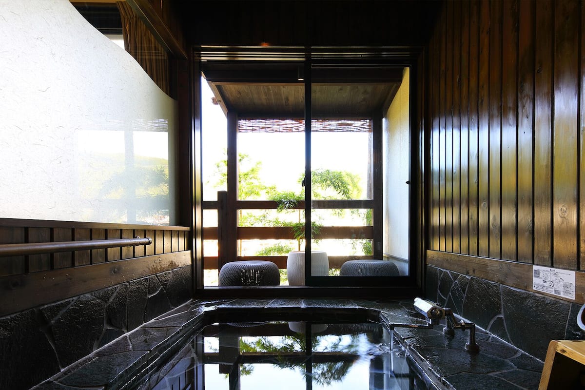Un jacuzzi con azulejos de piedra, un banco de madera, un taburete pequeño y una mesa de madera con un cuenco.