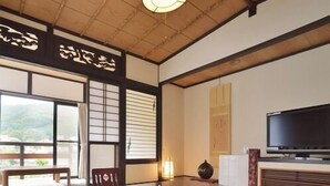 Ein traditioneller japanischer Raum mit Tatami-Bodenbelag, einem niedrigen Tisch mit Kissen, einem Fernseher und Schiebetüren.