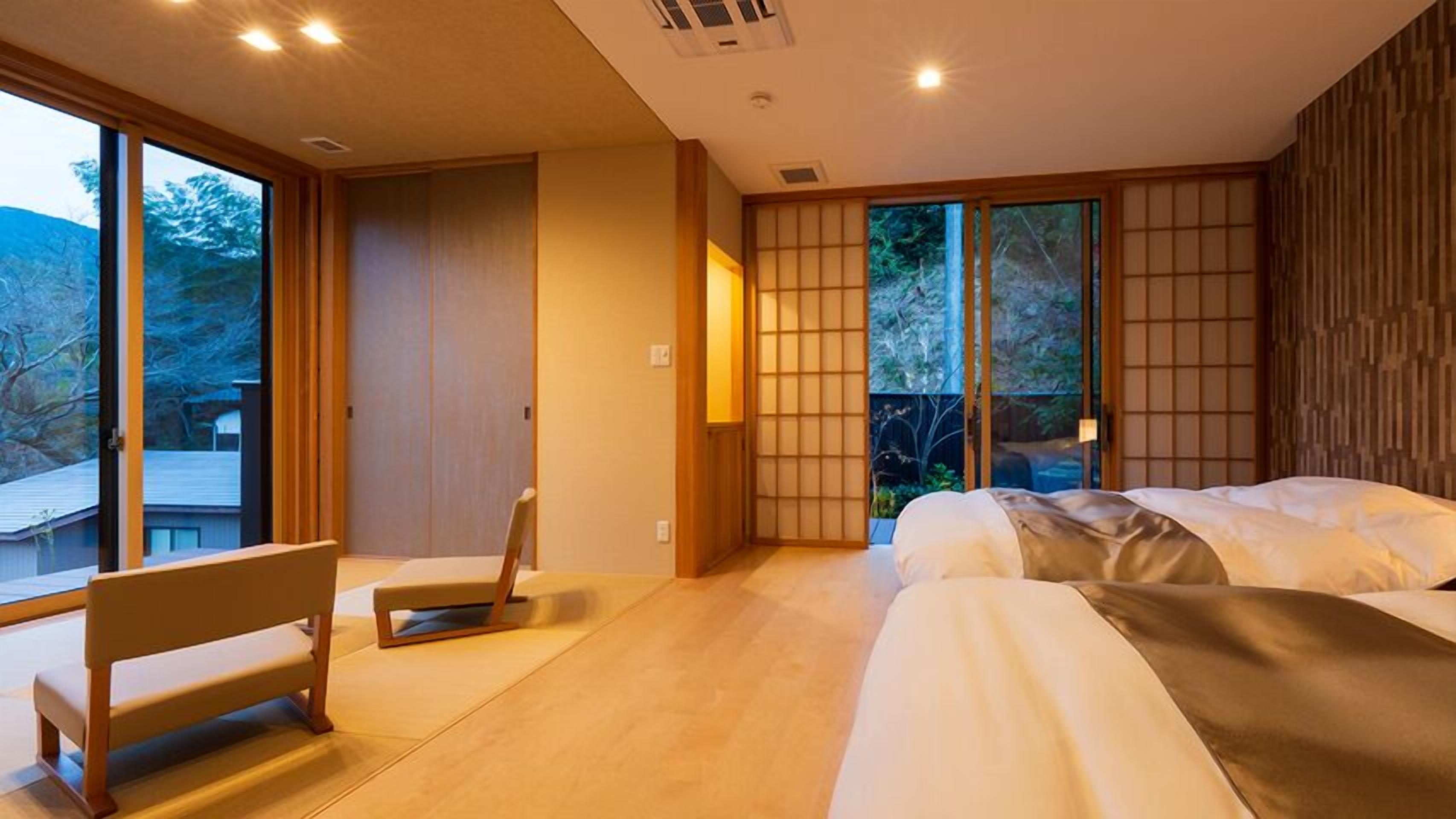 Una habitación de hotel moderna con un gran ventanal corredero, una cama, un banco y vistas al exterior.