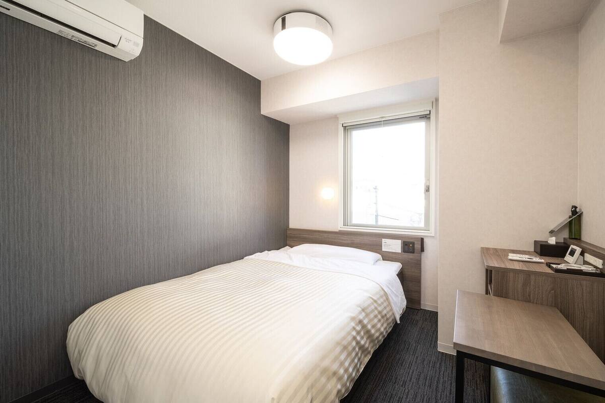 A hotel room with a bed, a desk, a chair, a window, and an air conditioning unit.