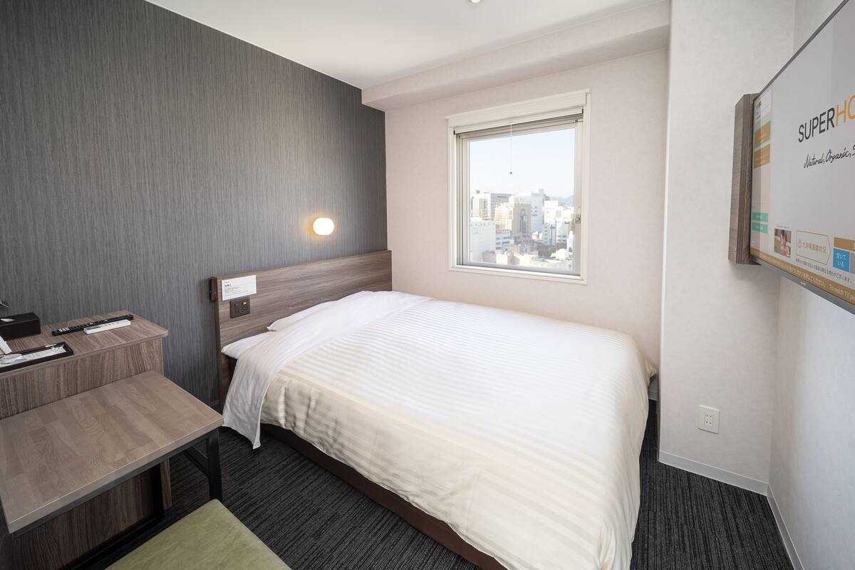 A hotel room with a bed, a desk, a window with a city view, and a wall-mounted TV.
