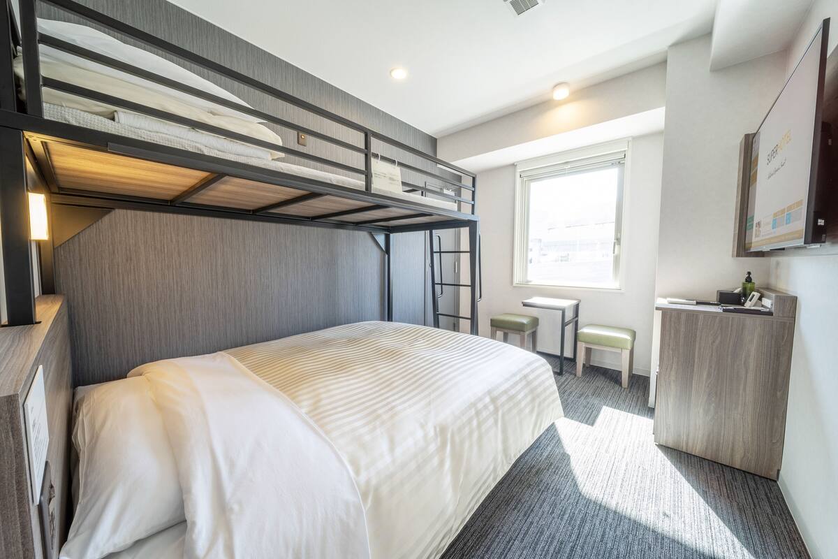 A hotel room with a bunk bed, a desk, a chair, and a window.