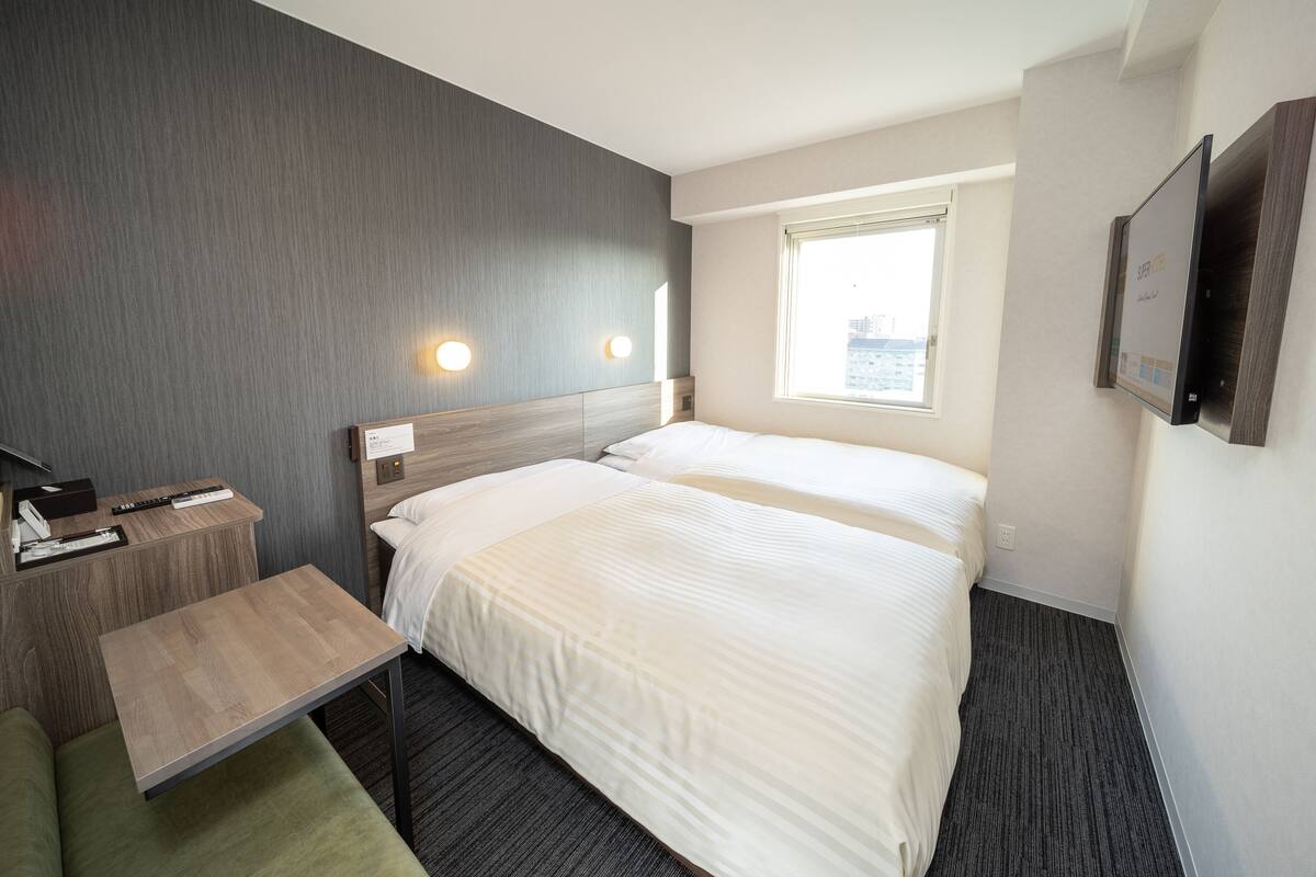 A hotel room with two beds, a desk, a chair, a TV, and a window with a city view.