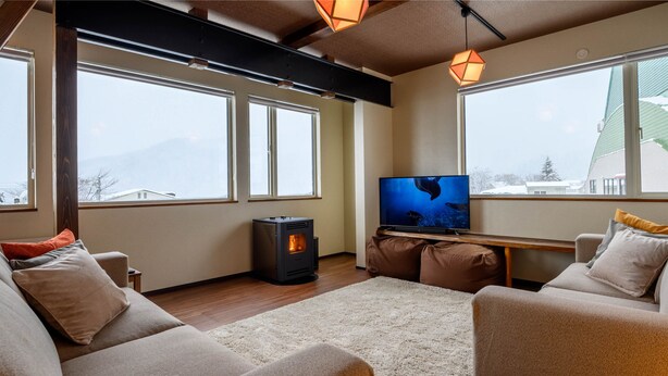 A cozy living room with a fireplace, comfortable seating, and large windows offering a view of the outdoors. - Iroha Nozawa (Nozawaonsen)