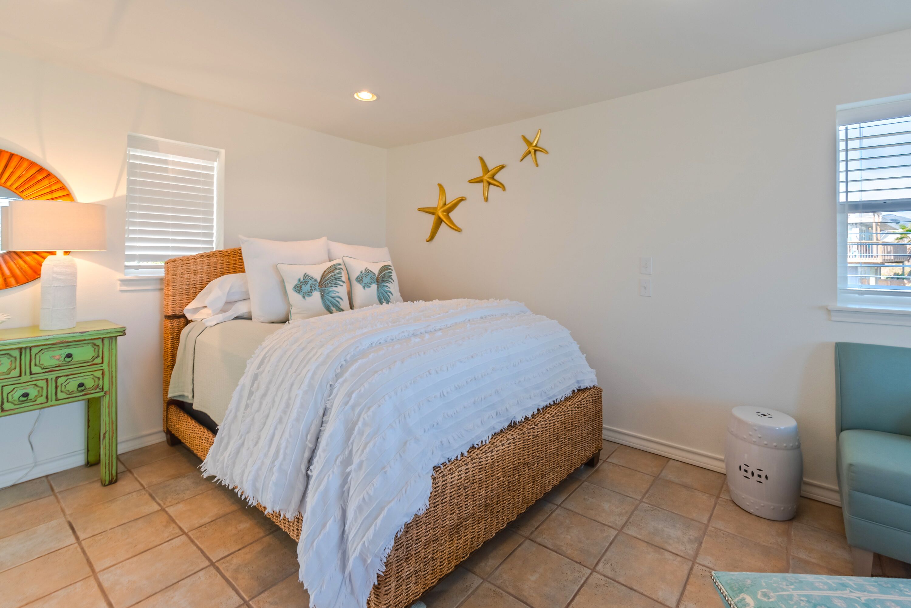 A bedroom with a wicker bed, white bedding, a green nightstand, a lamp, and a window with blinds.