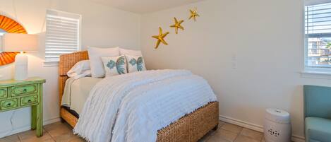 A bedroom with a wicker bed, white bedding, a green nightstand, a lamp, and a window with blinds.