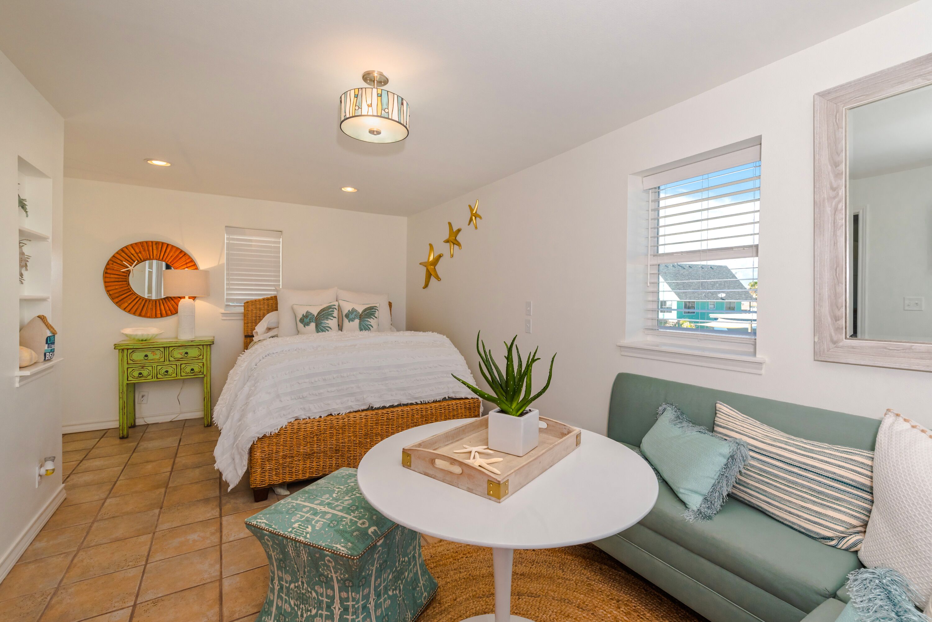 A bedroom with a bed, a green sofa, a round table, a green nightstand, a mirror, and a window with blinds.