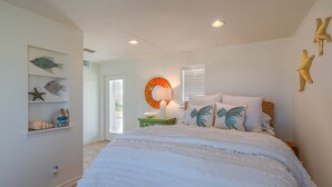 A bedroom with a large bed, a wall-mounted shelf with decorative items, a round mirror, and a window with blinds. - Bikini Bottom 2Bed 2Bath Ocean View Huge Outdoor Pool - DOWNSTAIRS UNIT (Port Aransas)
