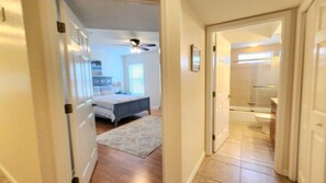 Un couloir mène à une chambre comprenant un lit, un ventilateur de plafond et une salle de bain avec des toilettes et une douche.