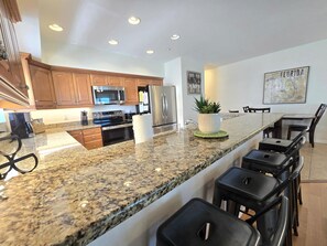 A modern kitchen with granite countertops, stainless steel appliances, wooden cabinets, and a dining area with bar stools. - Ocean Side 306 Corner Unit Beach front (Indian Rocks Beach)