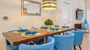 A dining area with a wooden table set for six, blue upholstered chairs, a hanging light fixture, a potted plant, and a mirror on the wall.