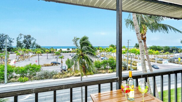 Een balkon met uitzicht op een parkeerplaats en strand, een fles alcohol en een glas op een houten tafel.