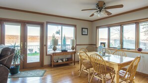 Un espace repas comprenant une table et des chaises en bois, un ventilateur de plafond et de grandes fenêtres donnant sur l’extérieur.