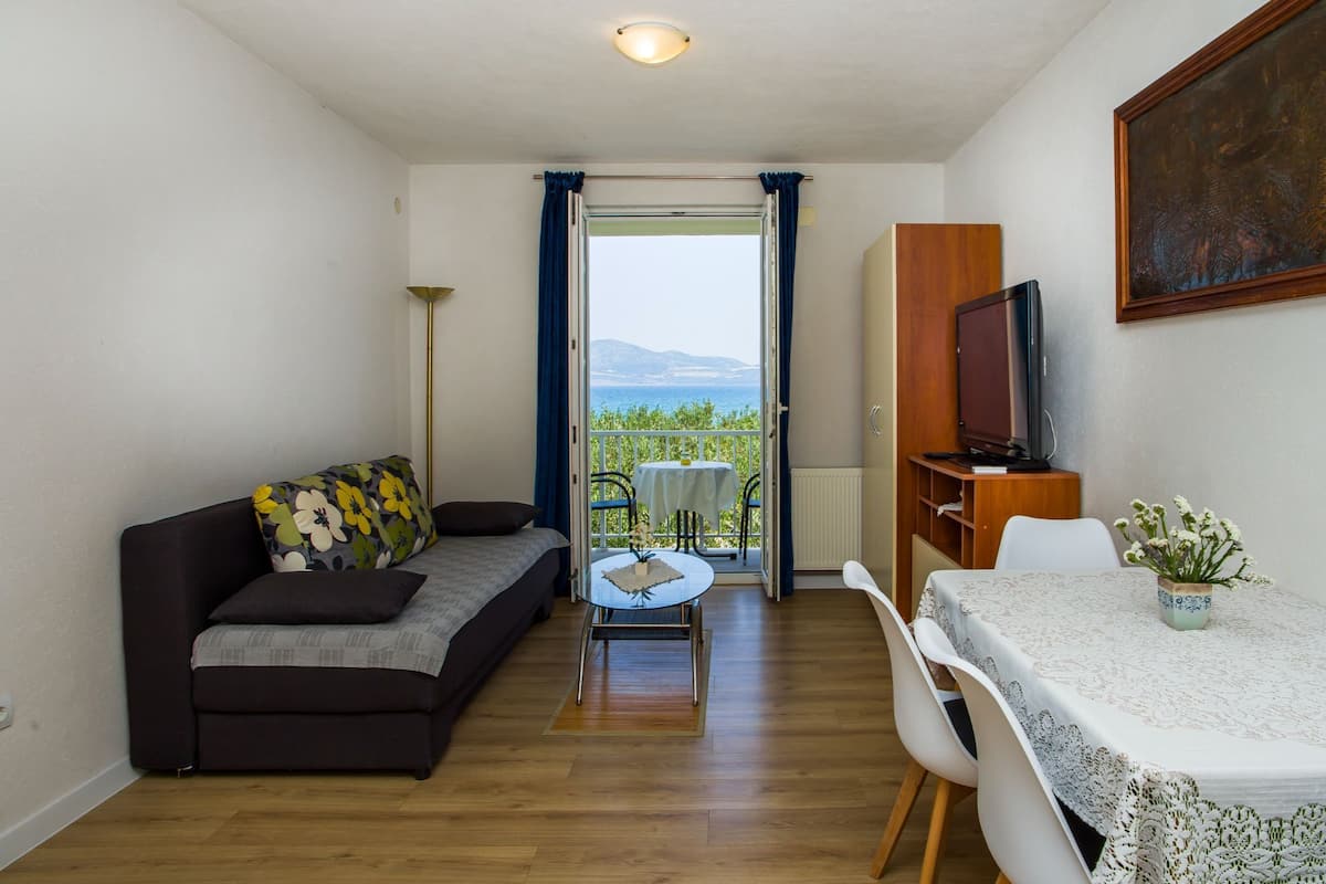 Una sala de estar compacta con un sofá, una mesa de comedor y un televisor. Se ve un balcón con vistas al mar a través de una puerta abierta.