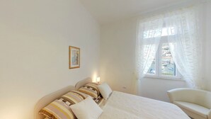 A bedroom with a bed, a chair, a nightstand, a lamp, and a window with curtains. - Apartment Mihe (Dubrovnik)