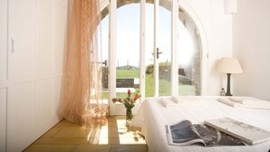 Een slaapkamer met een bed, een nachtkastje met een lamp, een raam met uitzicht op de omgeving, en een deur naar een balkon.