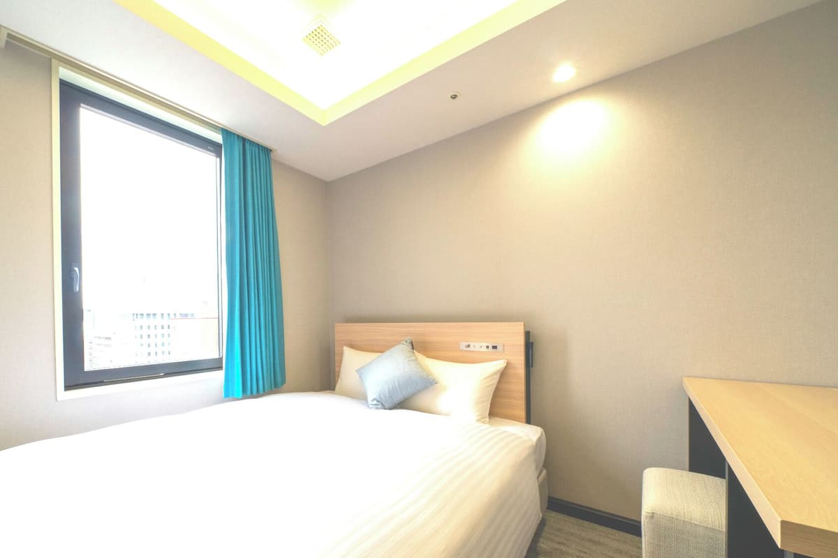 Une chambre d’hôtel avec un lit, un bureau, une fenêtre avec des rideaux et un luminaire au plafond.