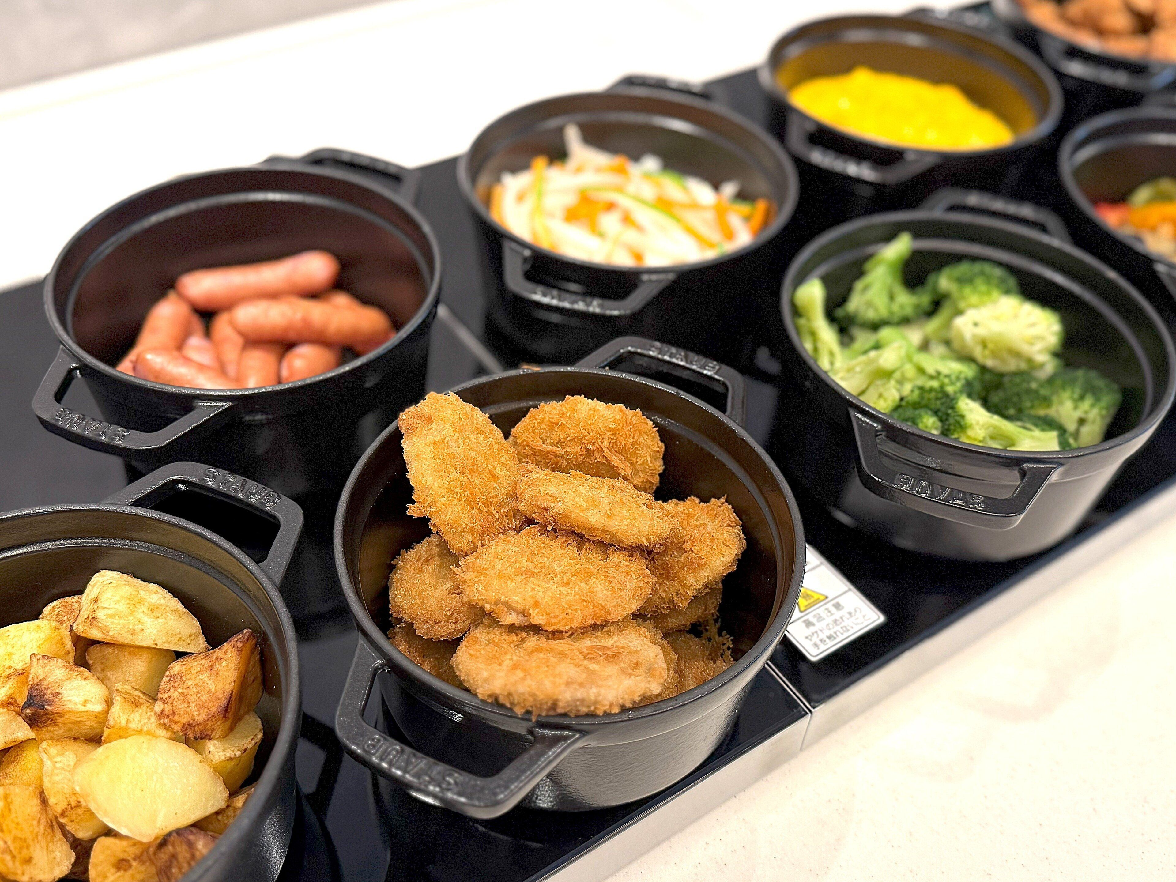 Un bufé con diversos platos, incluyendo salchichas, pollo frito, papas y verduras.