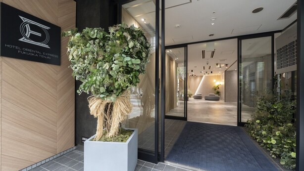 Ein Hotel Eingang mit einem großen Blumentopf, einem Empfangstresen und einem modernen Innenraum, der durch die Glastüren sichtbar ist.