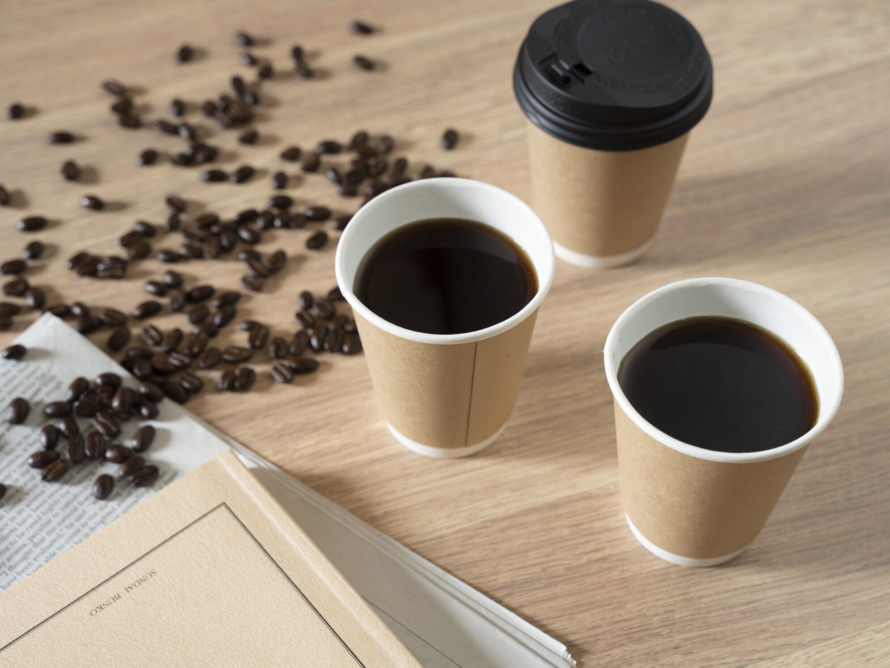 Three coffee cups on a wooden table with scattered coffee beans and a newspaper.