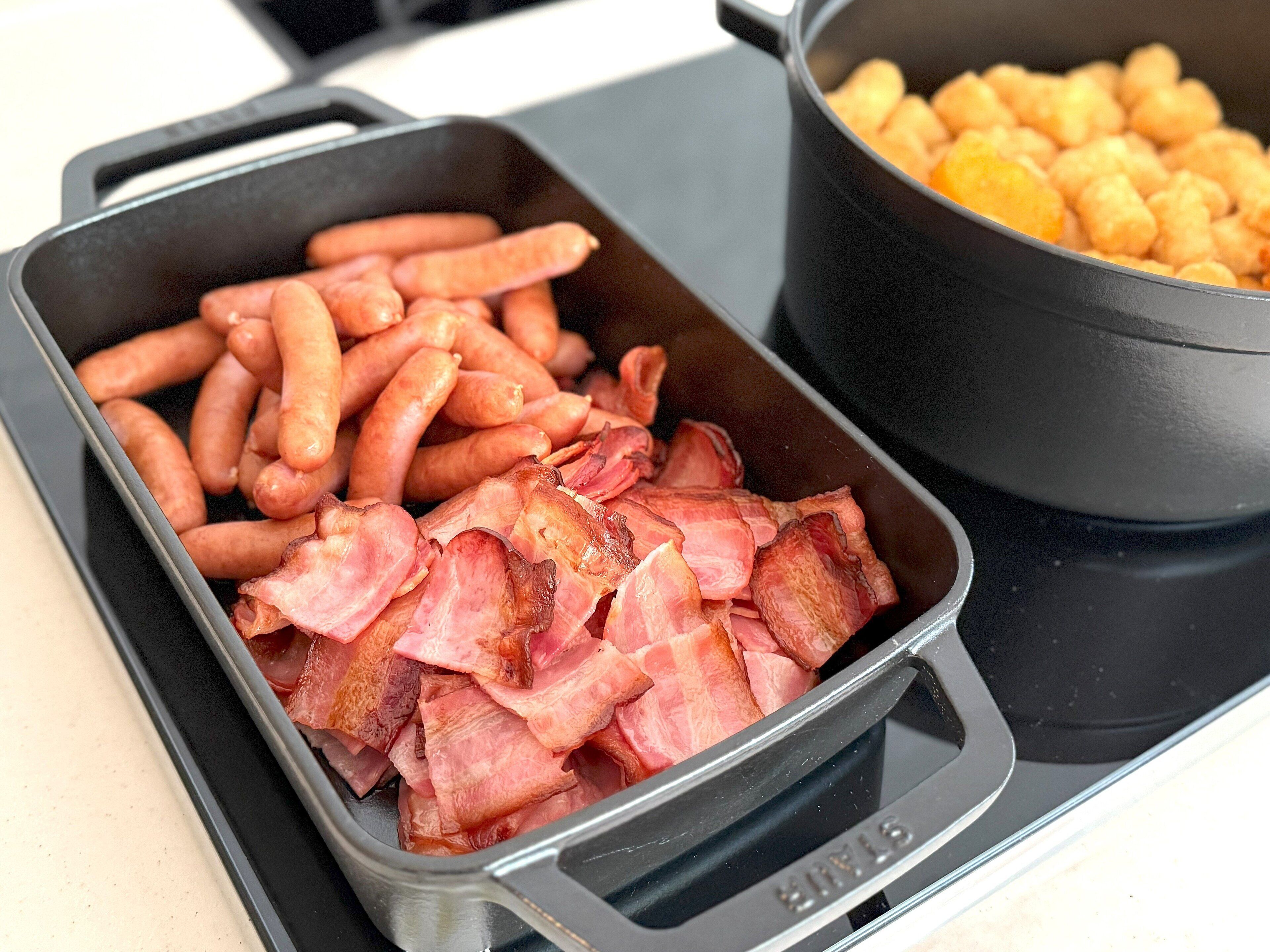 A kitchen with a black pot and a rectangular pan containing bacon and sausages.